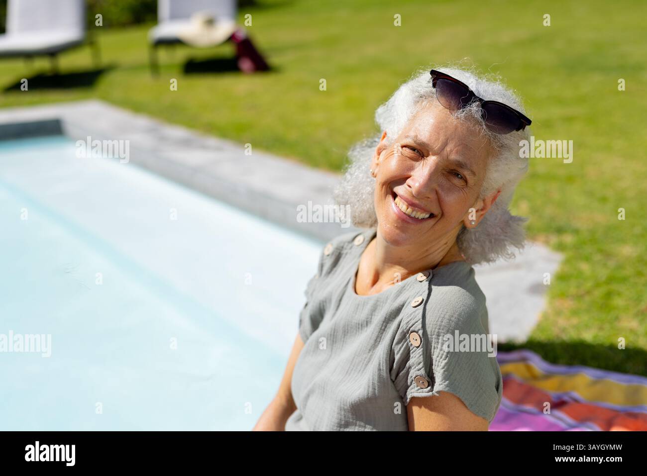 Femme senior assise au bord de la piscine dans la cour arrière, portant des lunettes de soleil et souriant sur une serviette, espace de copie Banque D'Images