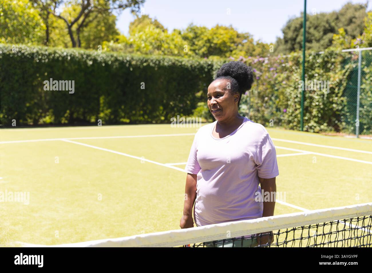 Femme afro-américaine senior debout et souriante sur un court de tennis, avec filet de tennis, lignes de court Banque D'Images