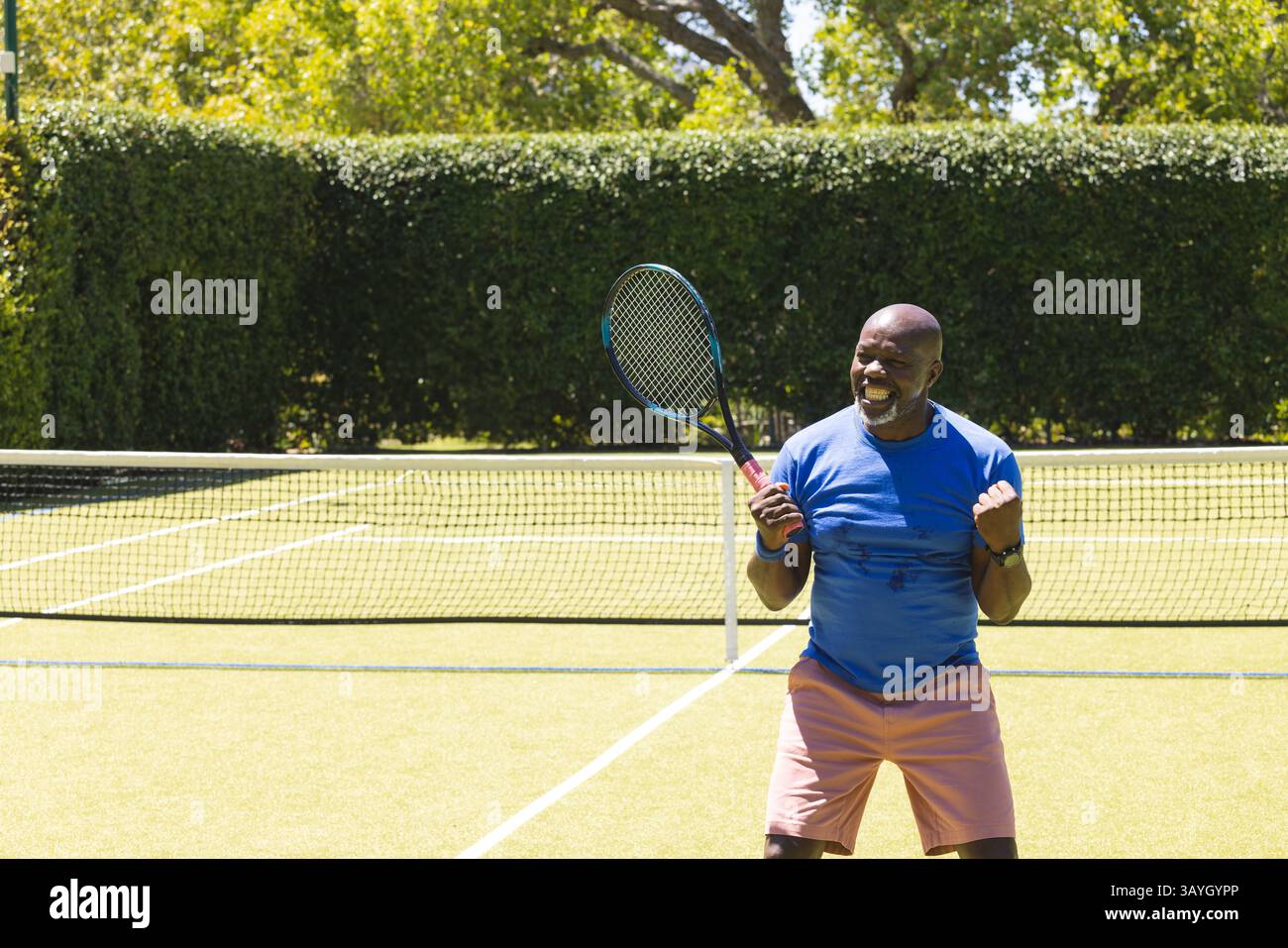 Homme afro-américain senior pompant le poing, tenant une raquette de tennis sur un court de tennis extérieur, espace copie Banque D'Images