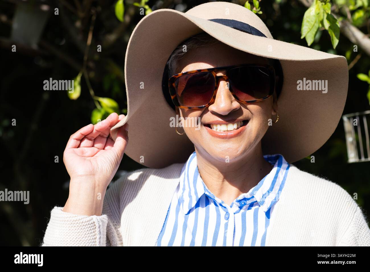 Femme souriante ajustant chapeau de soleil à large bord dans le jardin avec mangeoire en métal derrière, portant des lunettes de soleil Banque D'Images