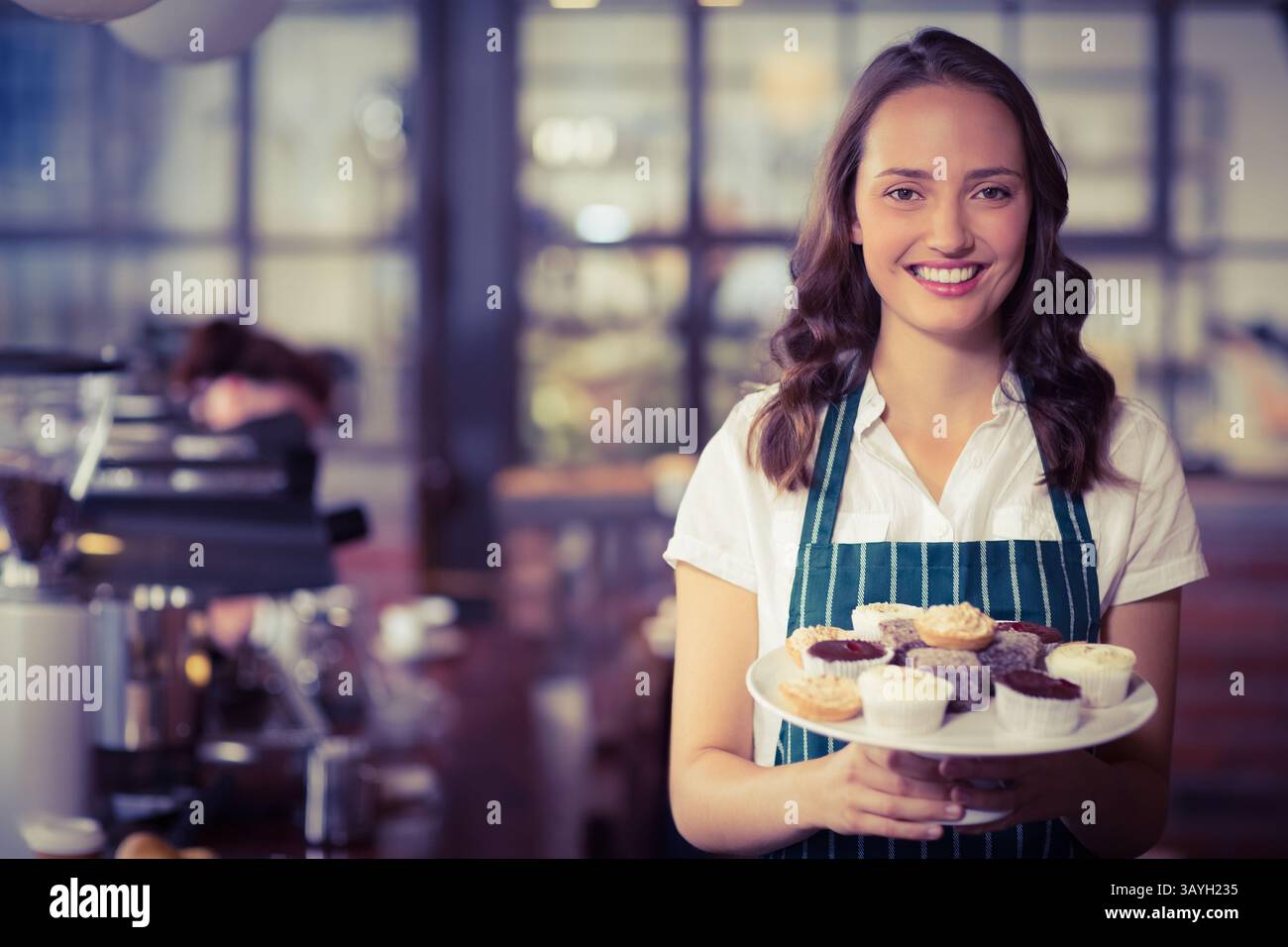 Jeune barista femelle tenant une assiette de cupcakes mélangés dans un café avec machine à expresso, espace de copie Banque D'Images