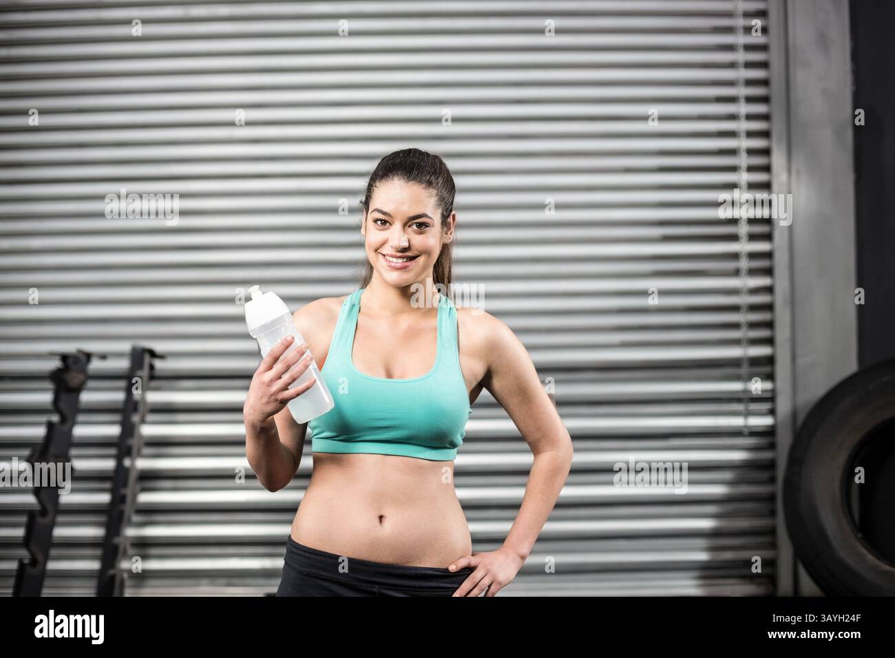 Femme debout et souriante à la salle de gym, tenant une bouteille de shaker blanche avec porte-poids, grand pneu de tracteur Banque D'Images