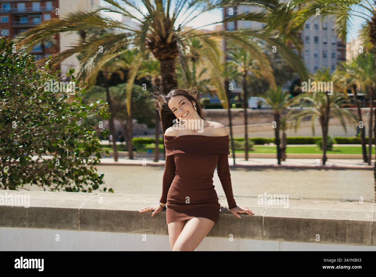 Heureuse jeune femme portant une couleur moka sur l'épaule mini robe souriante dans un parc avec des palmiers sur une journée ensoleillée, profitant des vacances d'été dans un Banque D'Images