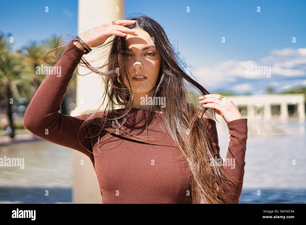 Jeune femme avec de longs cheveux bruns moka, portant une robe brune et des bijoux en or, protégeant ses yeux du soleil dans un parc de la ville avec un étang et des colonnes Banque D'Images
