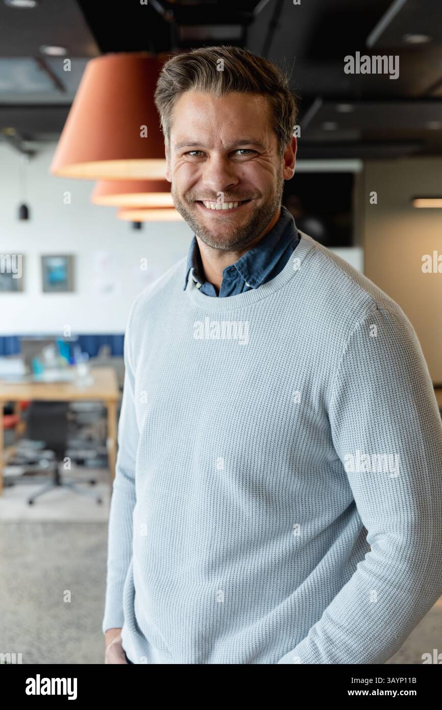Homme mi-adulte debout et souriant dans un bureau moderne ouvert, avec des bureaux en bois et des lampes suspendues Banque D'Images