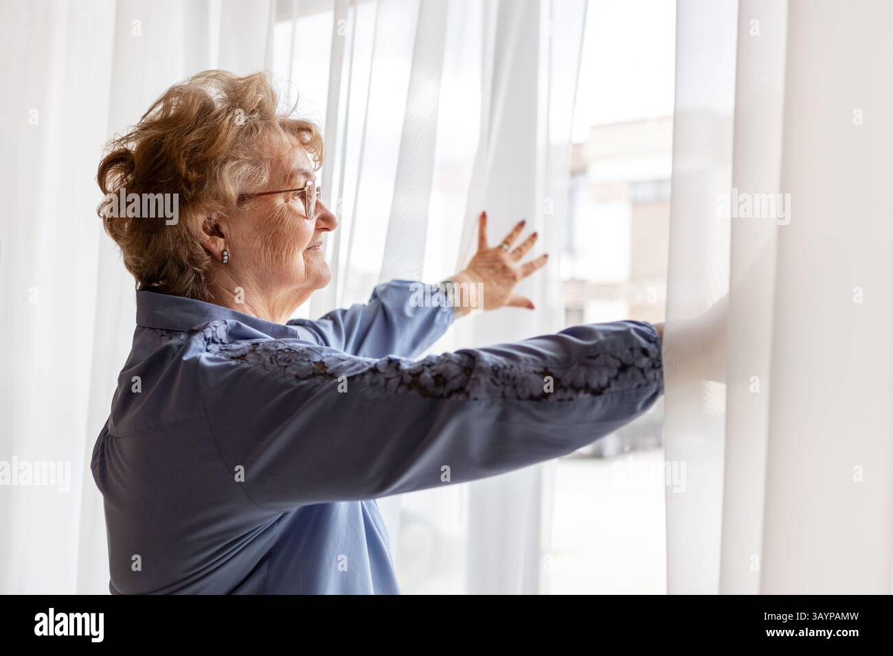 Femme âgée pairs espérons à travers de purs rideaux dans sa maison pendant la matinée. Humeur nostalgique, scène de confort à la maison pour le concept de style de vie de retraite a Banque D'Images