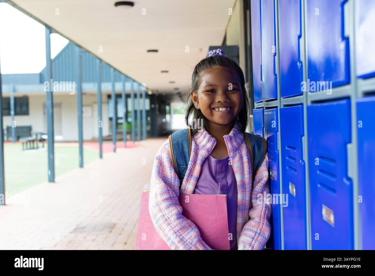 Fille multiraciale appuyée contre des casiers bleus dans la salle de l'école avec classeur et sac à dos, espace de copie Banque D'Images