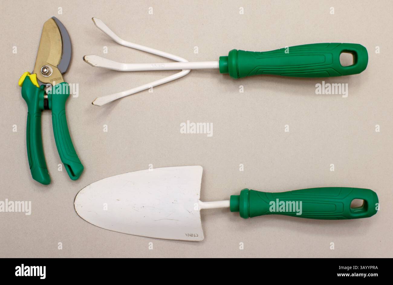 Trois outils de jardinage blancs et verts - une truelle, un râteau à main et un sécateur - sur un fond gris clair / brun. Banque D'Images