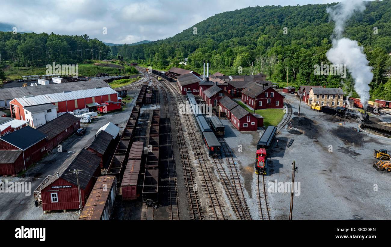 Une gare ferroviaire bien entretenue comprend des trains anciens stationnés à côté de bâtiments rustiques rouges. Des collines verdoyantes entourent la région, avec de la vapeur bouillonnante Banque D'Images