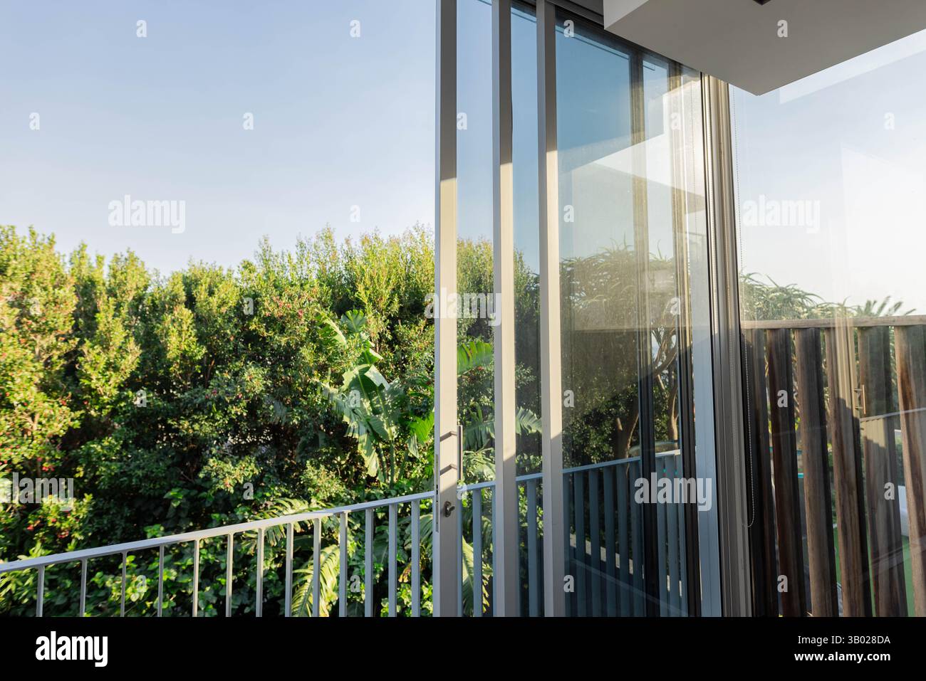 La porte vitrée coulissante révèle la balustrade du balcon tandis que la lumière du soleil met en évidence la haie verte à l'extérieur Banque D'Images