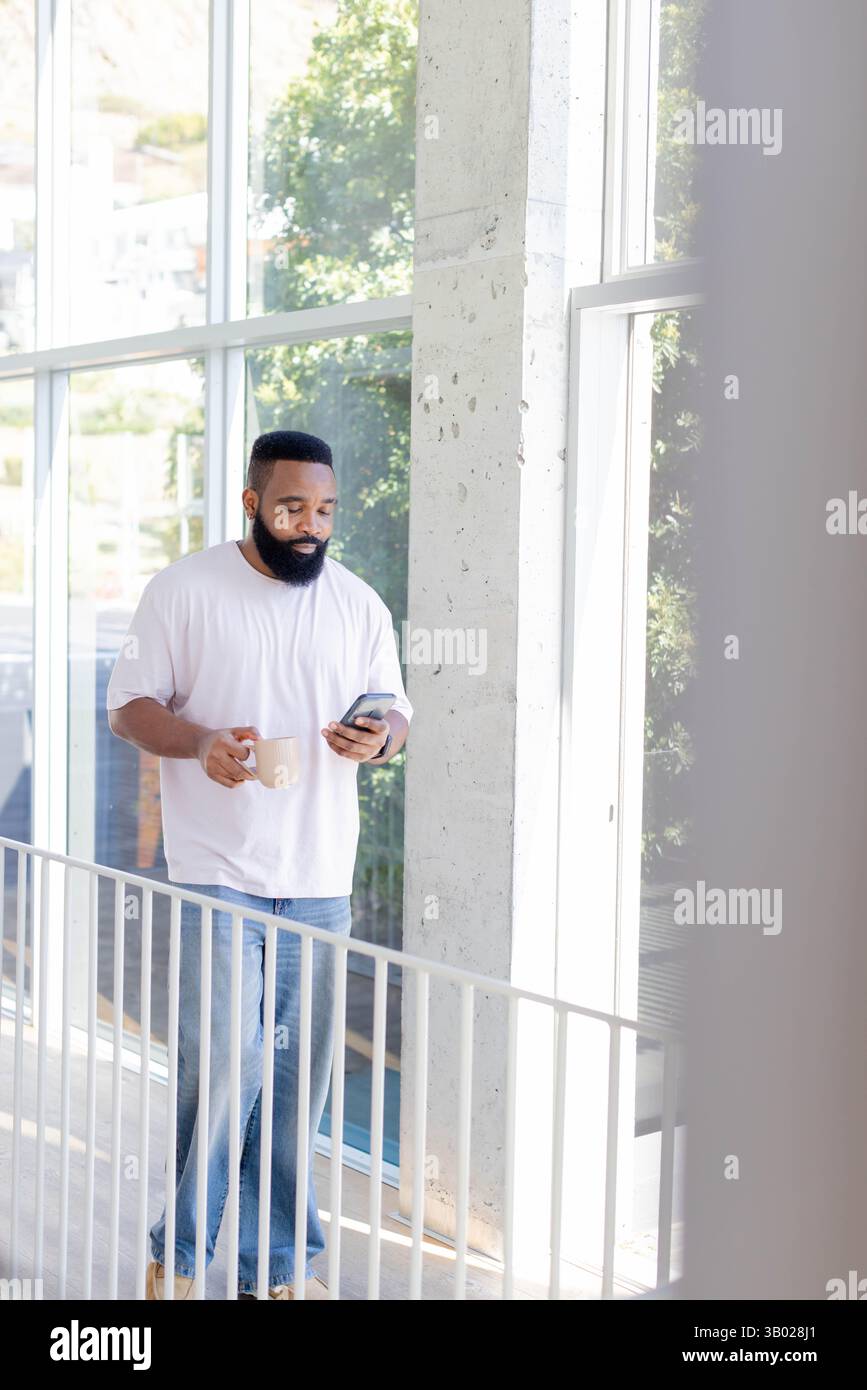Homme afro-américain marchant tenant une tasse de café à l'aide d'un smartphone sur la balustrade dans le couloir, espace de copie Banque D'Images