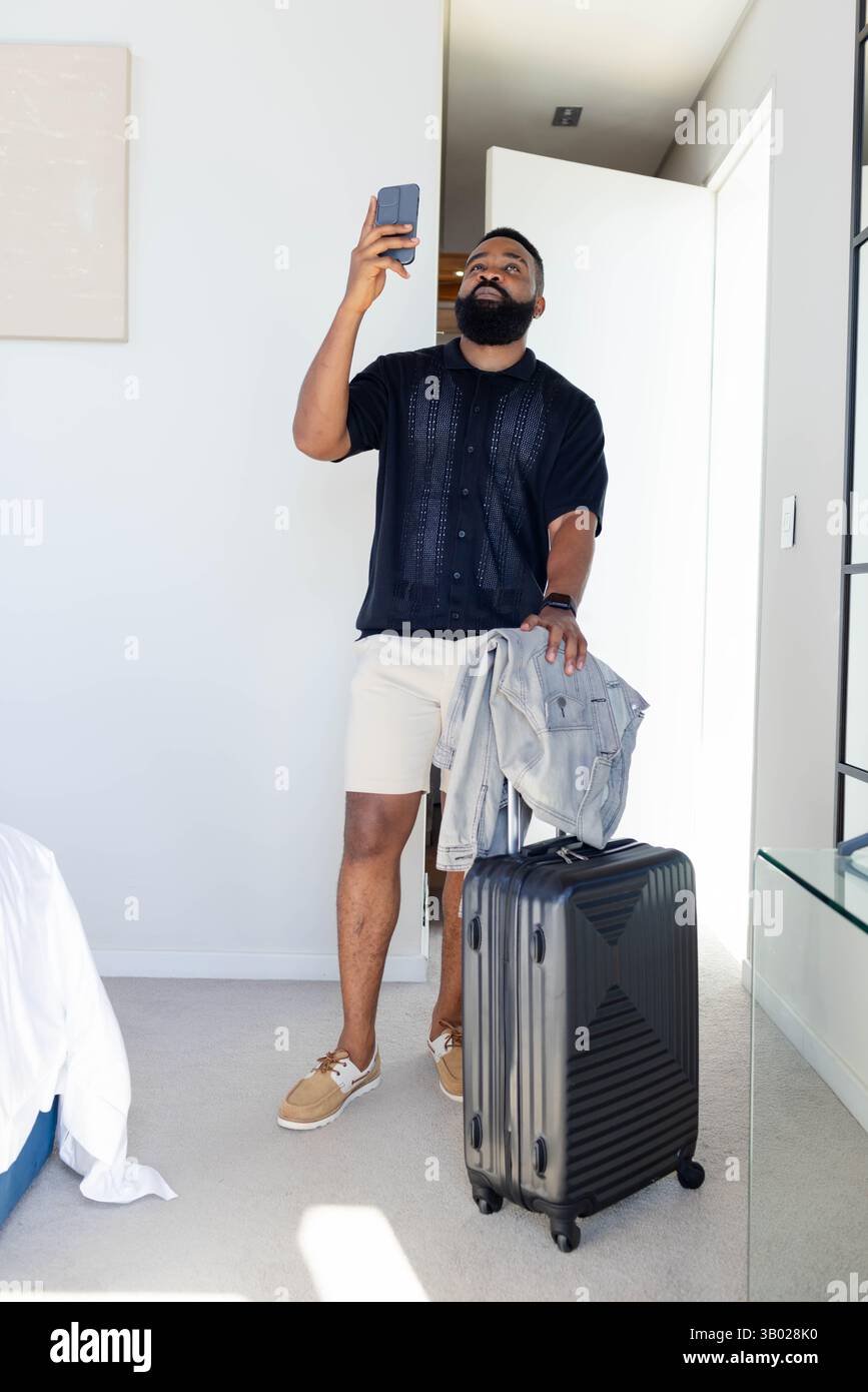 Homme afro-américain debout tenant smartphone par porte ouverte dans la chambre d'hôtel, avec valise Banque D'Images
