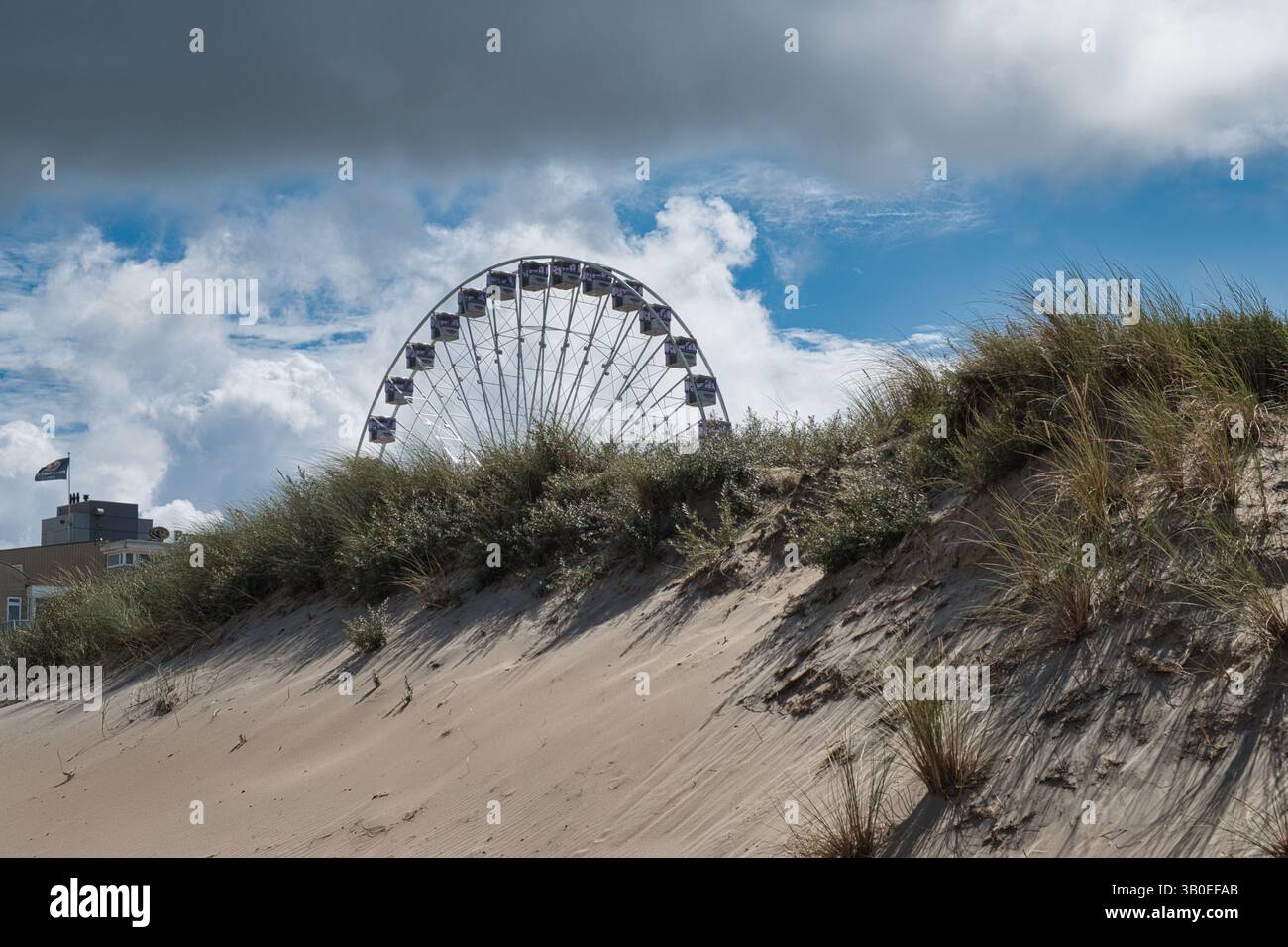 Une grande roue émerge derrière de douces dunes de sable avec de l'herbe, encadrée par des nuages et un ciel bleu dans un paysage côtier. Banque D'Images