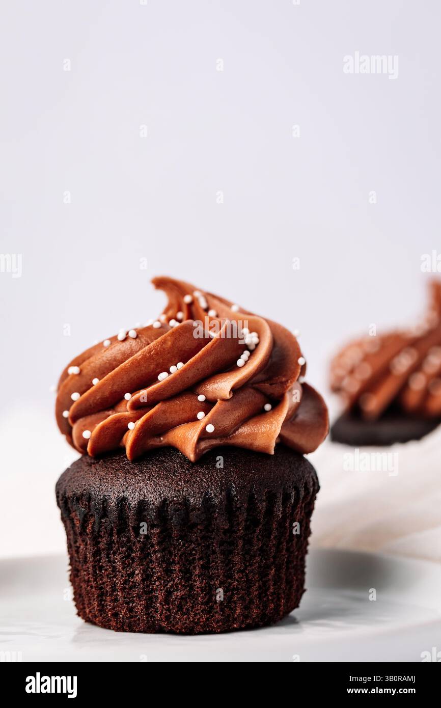 Cupcake au chocolat recouvert de glaçage au chocolat tourbillonnant et de petites saupoudrées blanches sur un fond blanc minimaliste. Banque D'Images