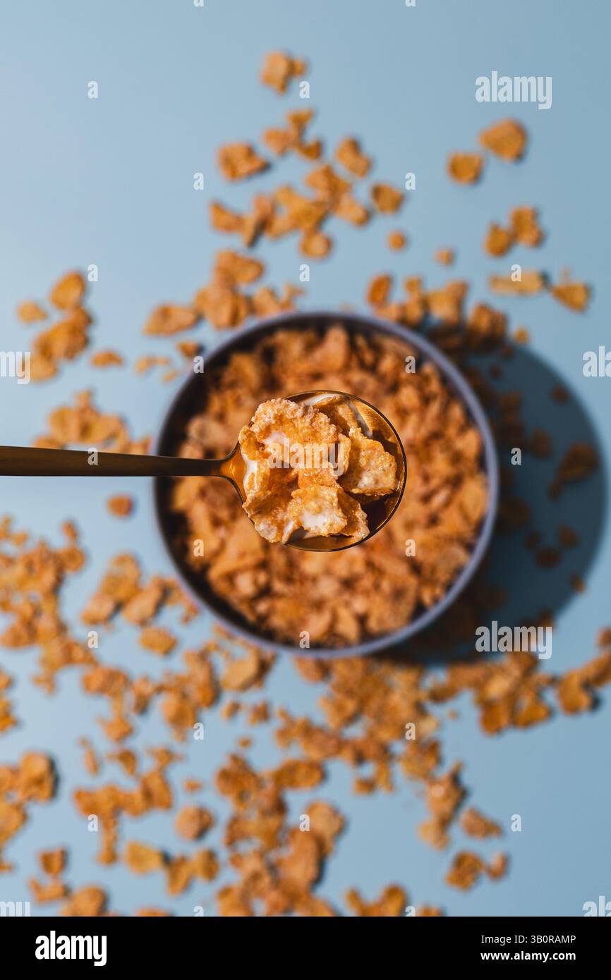 Une cuillerée de cornflakes dorés au lait planent au-dessus d'un bol de céréales, entouré de flocons dispersés sur un fond bleu audacieux - ludique et moderne. Banque D'Images Une cuillerée de cornflakes dorés au lait planent au-dessus d'un bol de céréales, entouré de flocons dispersés sur un fond bleu audacieux - ludique et moderne. Banque D'Images