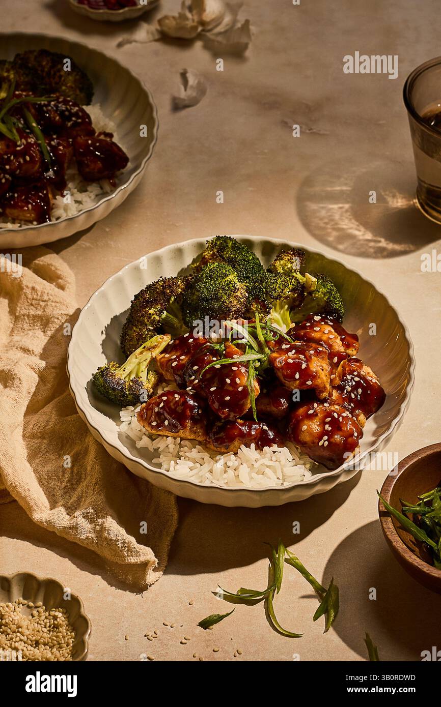Un bol de poulet du général Tso avec du brocoli rôti et du riz blanc, garni de graines de sésame et d’oignons verts, décoré dans un éclairage chaud et dramatique. Banque D'Images Un bol de poulet du général Tso avec du brocoli rôti et du riz blanc, garni de graines de sésame et d’oignons verts, décoré dans un éclairage chaud et dramatique. Banque D'Images