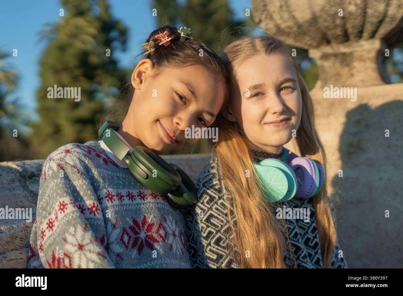 Deux filles portant des écouteurs autour du cou posent ensemble dans un parc, profitant du soleil et appréciant la musique tout en partageant des rires et en créant des las Banque D'Images