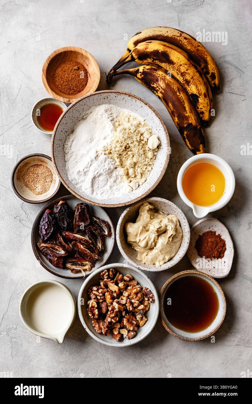 Assortiment d'ingrédients pour une recette comprenant des bananes mûres, diverses noix, de la farine et des épices disposées sur une surface texturée. Banque D'Images