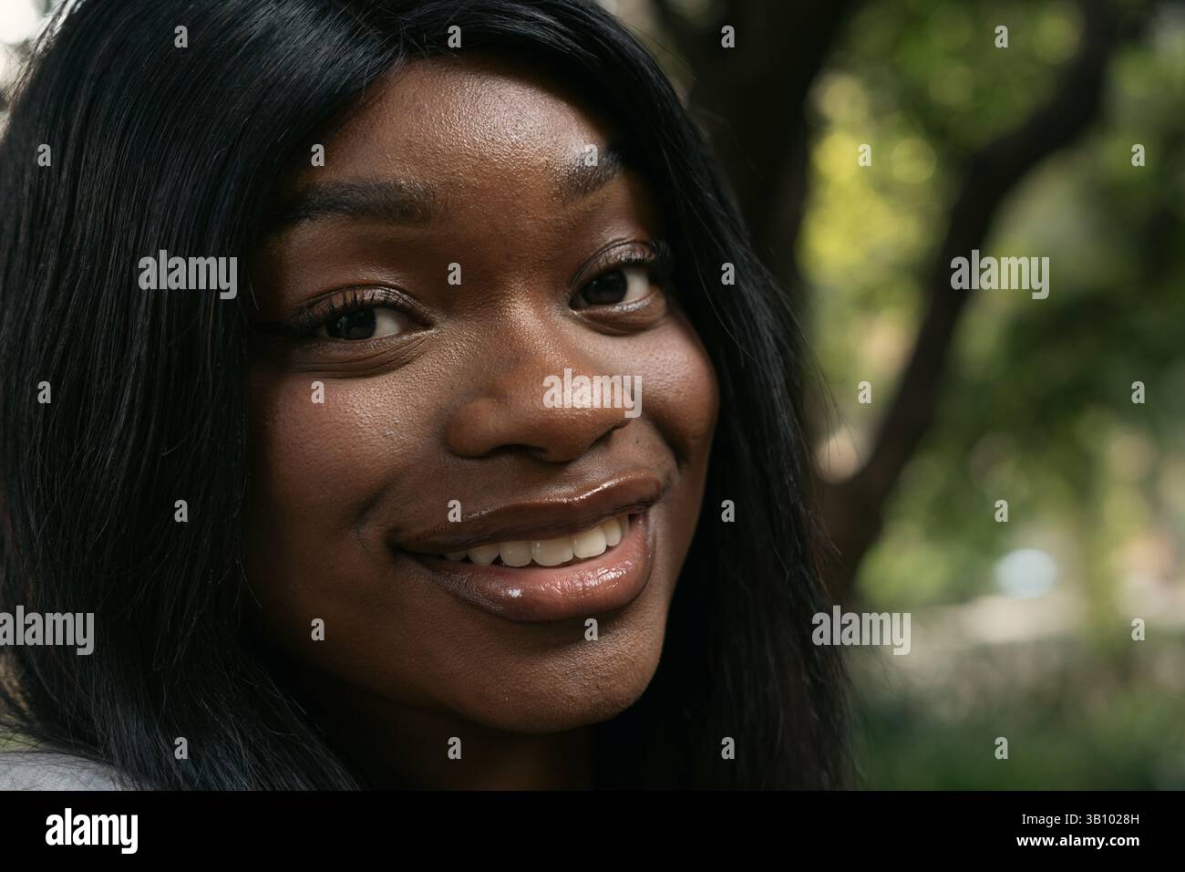 Portrait rapproché d'une belle jeune femme afro-américaine aux longs cheveux noirs souriants dans un parc, profitant d'une journée ensoleillée entourée par la nature Banque D'Images