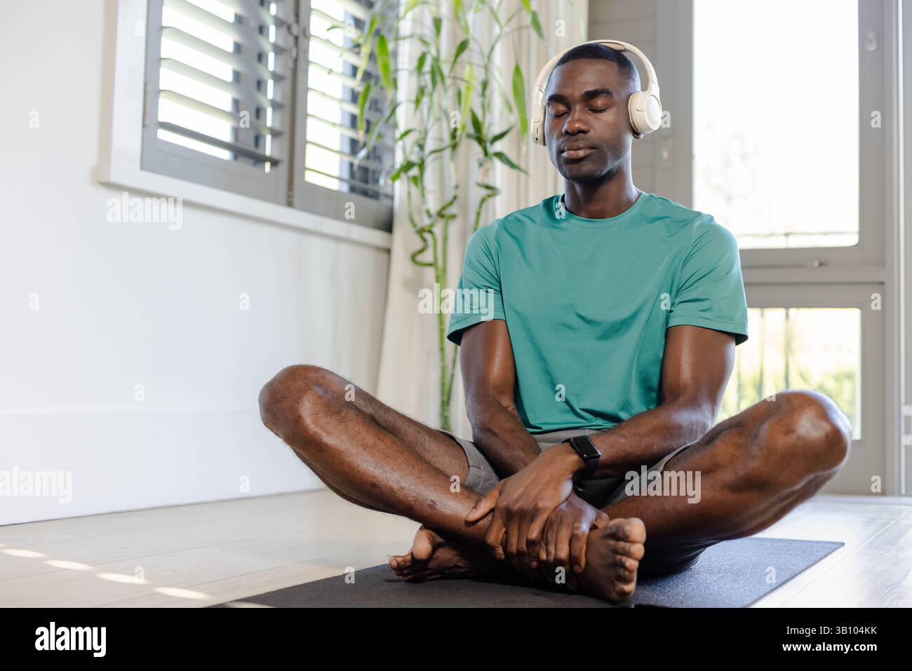 Homme afro-américain méditant assis les jambes croisées dans le coin de méditation avec tapis de yoga, écouteurs Banque D'Images
