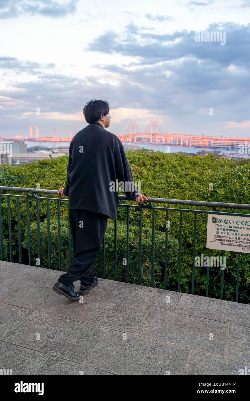 Un Japonais dans la vingtaine portant une veste noire se trouve à Minato Mirai, Yokohama, préfecture de Kanagawa. Vu de derrière, il regarde un pont lointain Banque D'Images