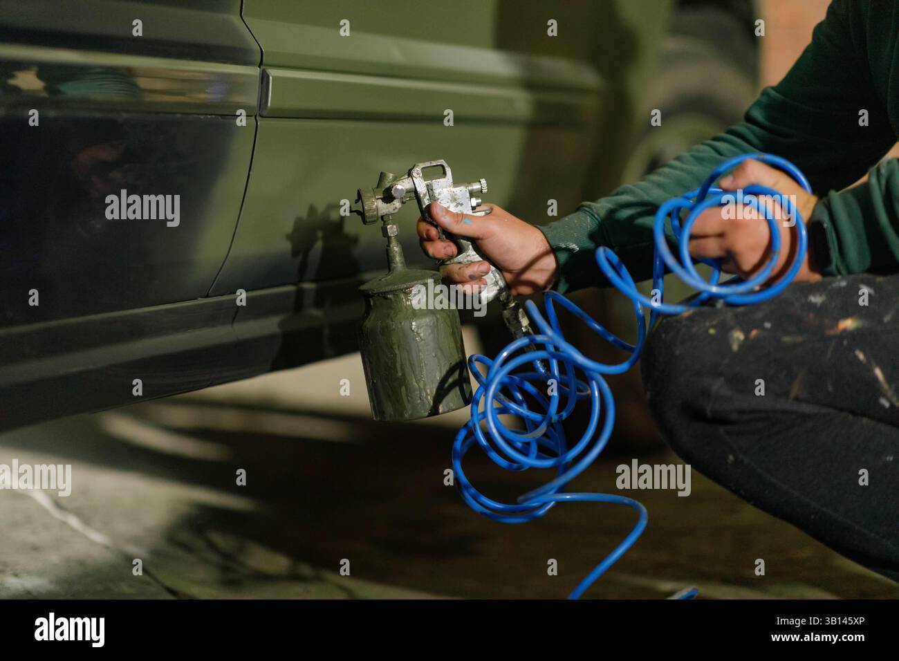 Une personne se concentre sur la peinture d'une voiture avec un pistolet de pulvérisation tout en étant assis sur le plancher d'un garage. L'environnement est faiblement éclairé, typique d'un pro du soir Banque D'Images