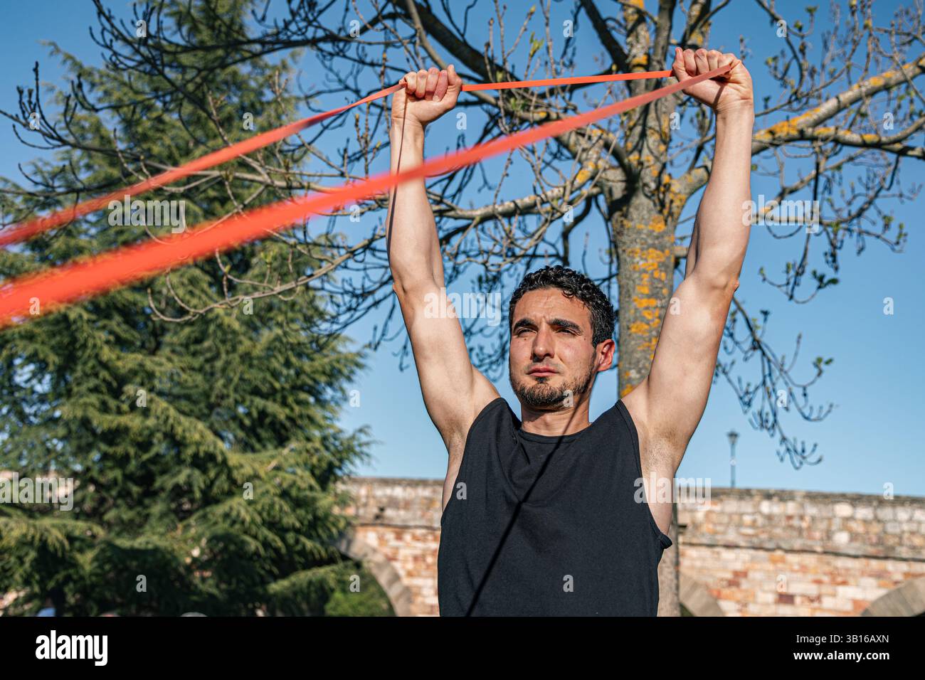 Cette image montre un jeune homme déterminé élevant une bande de résistance rouge au-dessus de sa tête pendant une séance d'entraînement en extérieur. Ses bras sont complètement étendus Banque D'Images
