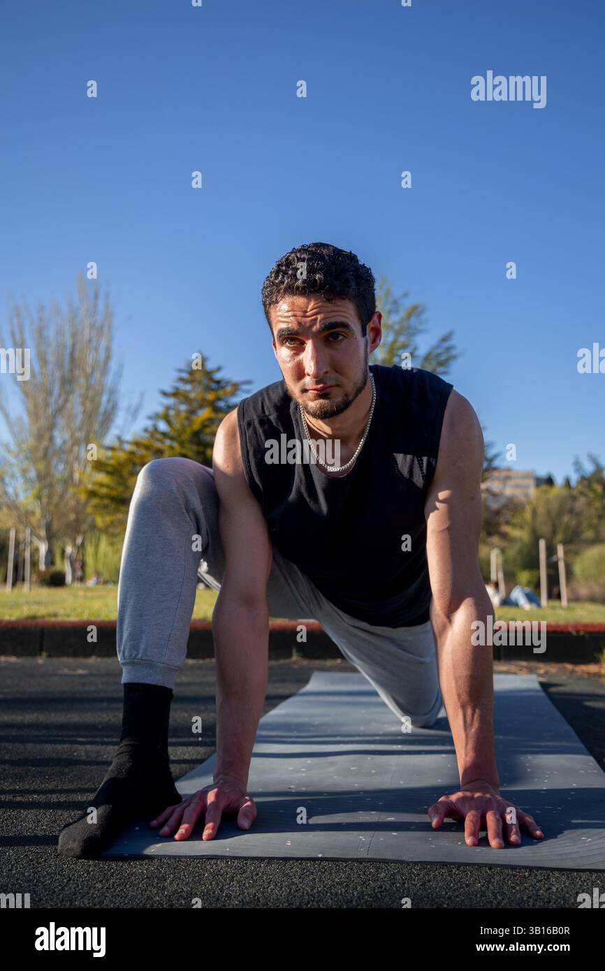 Homme athlétique s'étirant sur le tapis d'exercice dans le parc pendant la journée ensoleillée, se préparant à l'entraînement avec concentration et équilibre en position de fente basse Banque D'Images