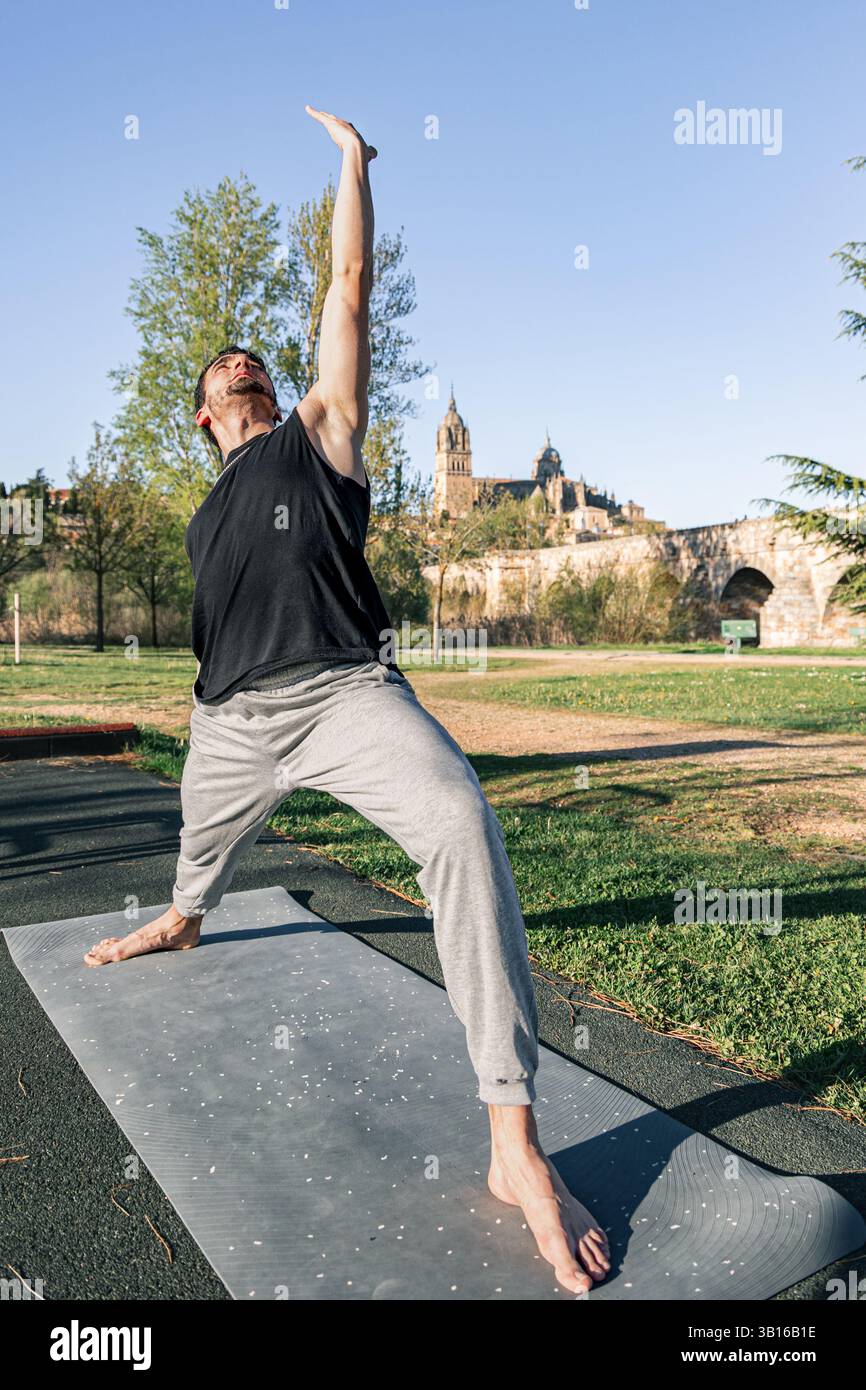 Homme pratiquant le yoga en guerrier inversé étendu pose sur tapis dans le parc avec l'architecture historique à Salamanque, Espagne sous ciel bleu clair Banque D'Images