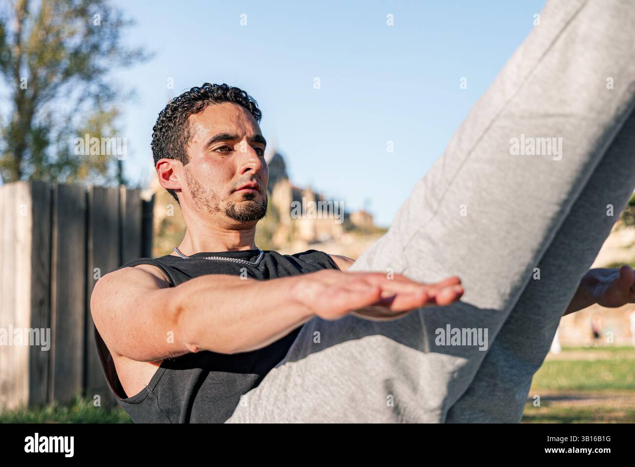 Homme concentré tenant la position de base V-SIT à l'extérieur par jour ensoleillé, mettant en valeur la force, l'équilibre et le contrôle du corps pendant la routine d'entraînement abdominale Banque D'Images