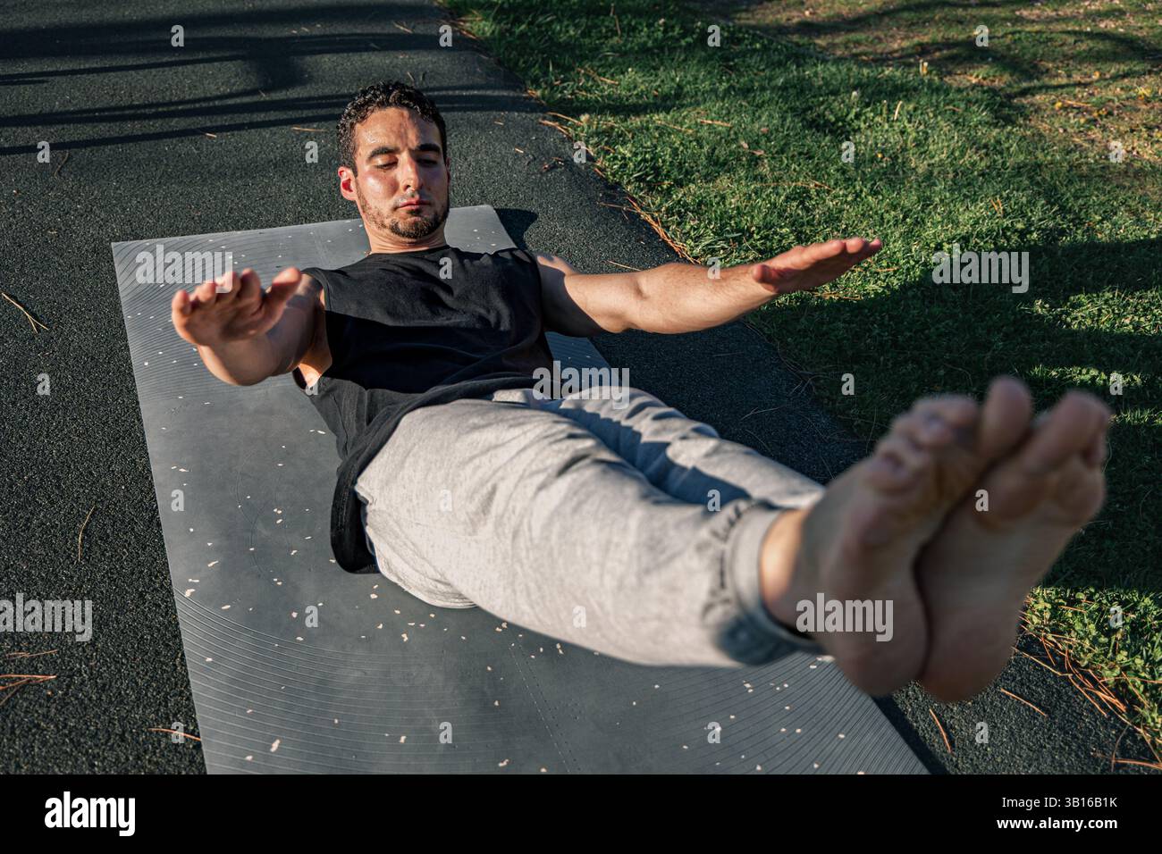 Jeune homme effectuant une prise de corps creuse sur un tapis extérieur avec les bras tendus et les jambes levées, montrant la force du noyau, l'équilibre et le contrôle sous la lumière du soleil Banque D'Images