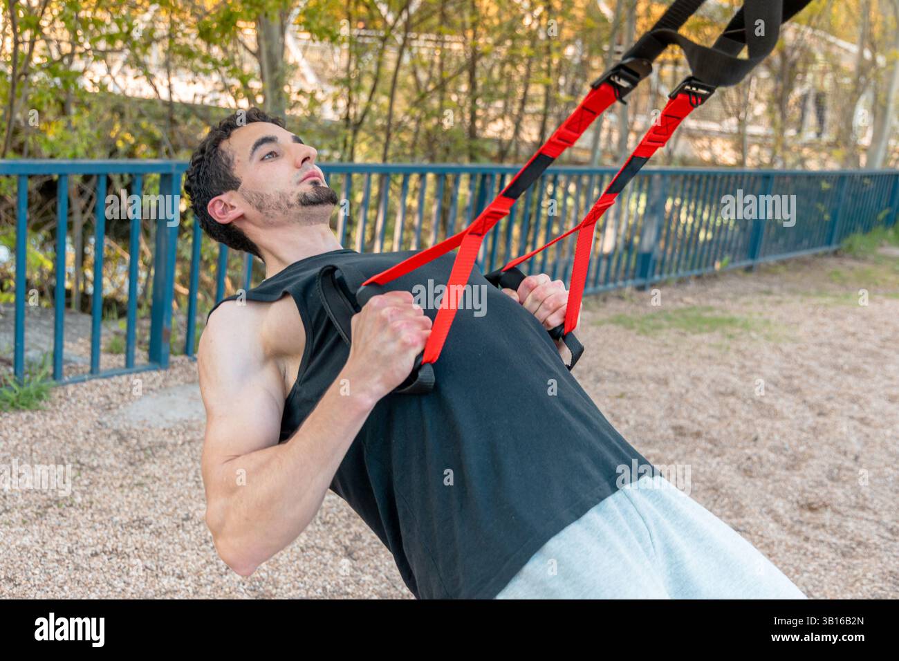 Homme athlétique penché dans la rangée TRX à l'extérieur sur une surface de gravier, tirant les sangles de suspension rouges à hauteur de poitrine avec une forme contrôlée et une tension musculaire Banque D'Images