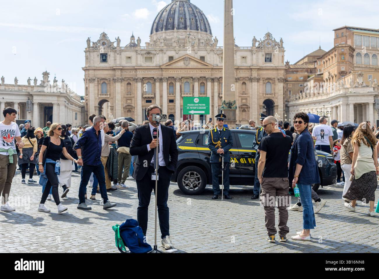 Vatican - 23 avril 2025 : journaliste reportant en direct de la place avec une foule de deuil en arrière-plan. Banque D'Images