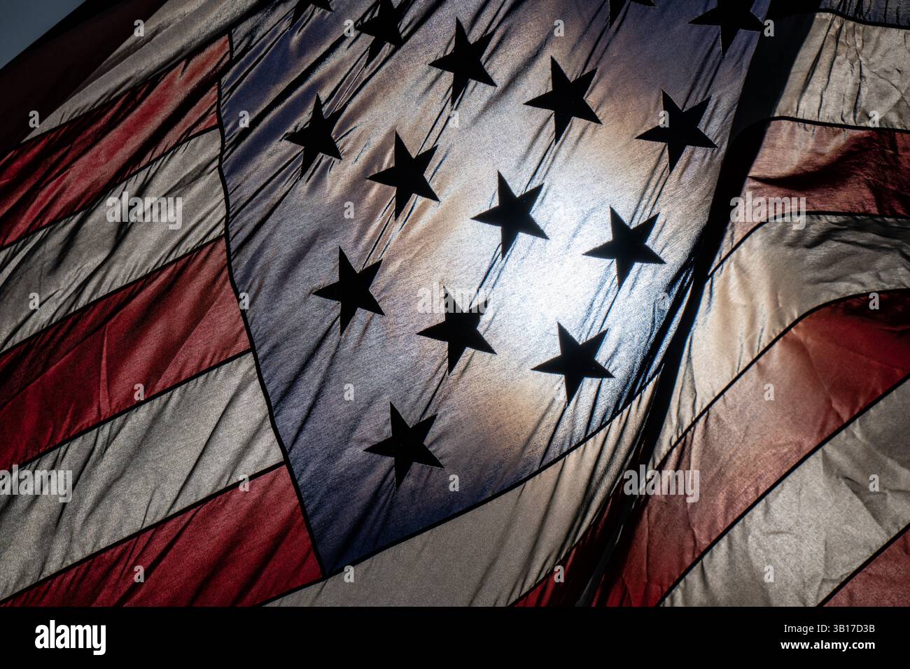 Drapeau des États-Unis flottant et éclairé par le soleil | ARLINGTON, Va. — La lumière du soleil perce le tissage du drapeau américain au Memorial de la Guerre du Corps des Marines, projetant ses étoiles en silhouette nette sur un champ de tissu lumineux. Le drapeau flotte continuellement au-dessus du mémorial selon une proclamation de 1961 du président Kennedy, un honneur accordé à un petit nombre de sites américains. Banque D'Images