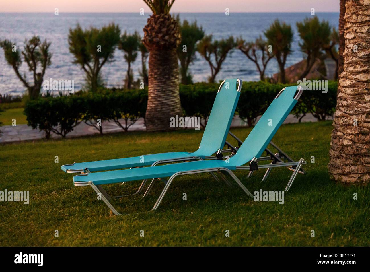 Deux chaises longues turquoise vides sont assises sur une pelouse herbeuse au bord de la mer au coucher du soleil, offrant un lieu de détente tranquille à la Canée, en Crète, en Grèce. Banque D'Images