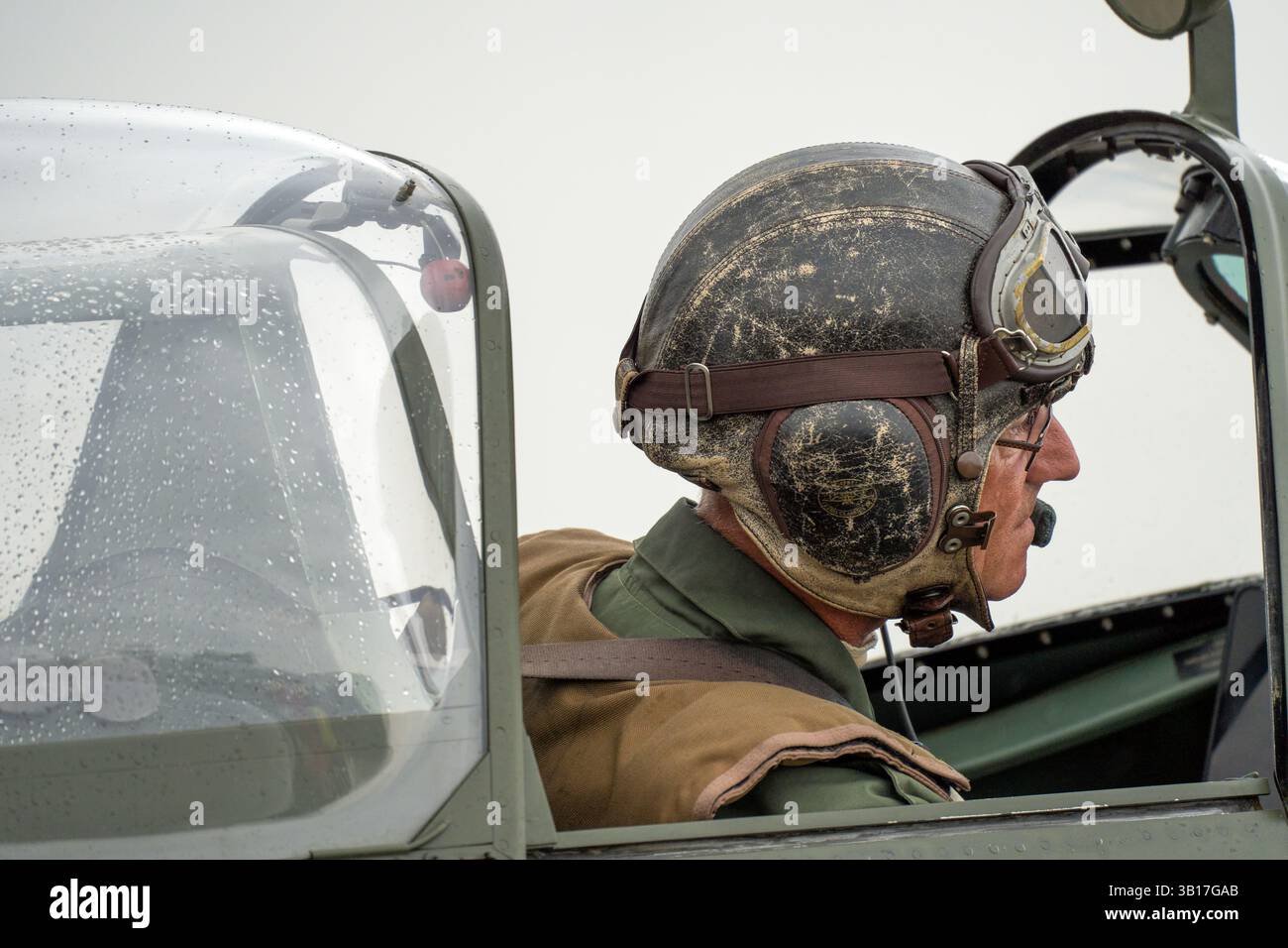 Pilote en cuir vintage casque et lunettes est assis dans le cockpit d'un Spitfire de la RAF, gouttelettes sur la canopée faisant allusion à la pluie récente à Duxford, en Angleterre. Banque D'Images