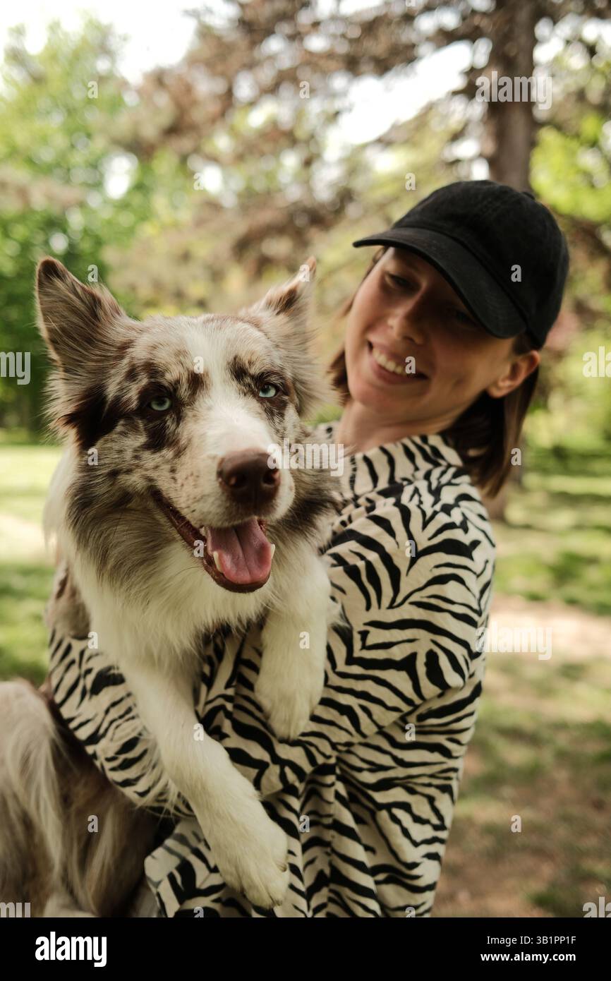 Heureuse jeune femme tenant un adorable chien rouge merle Border Collie dans ses bras, tous deux souriants dans un parc ensoleillé. Moment lumineux et joyeux plein d'amour et Banque D'Images