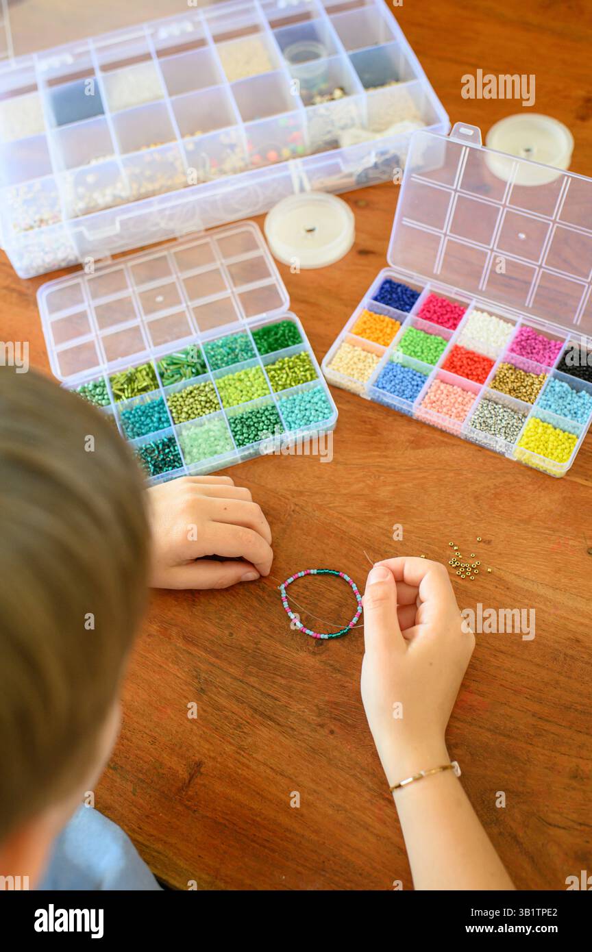 Un jeune garçon est assis à la maison devant de petits récipients remplis de perles colorées, les enfilant soigneusement pour faire un bracelet d'amitié fait à la main pour Banque D'Images