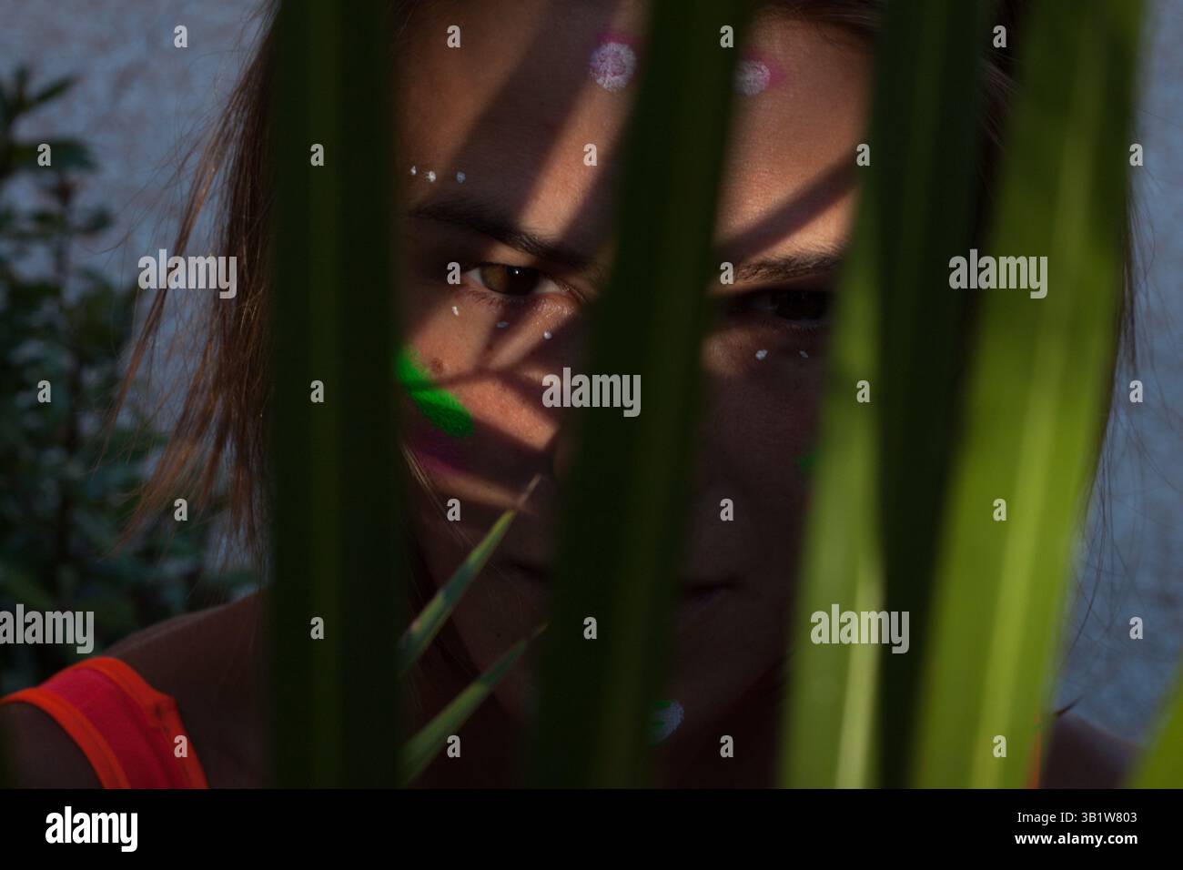 Gros plan détaillé des yeux de la belle femme avec la peinture créative au néon maquillage montrant sa partie de visage à travers des feuilles de palmier. Banque D'Images