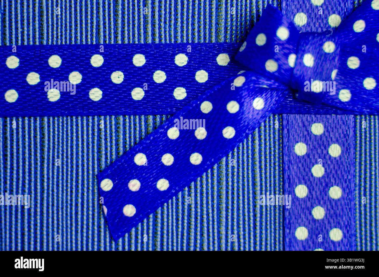 Gros plan d'un ruban cadeau bleu avec des pois blancs attachés dans un noeud, placé sur un fond rayé rouge et rose texturé. Banque D'Images