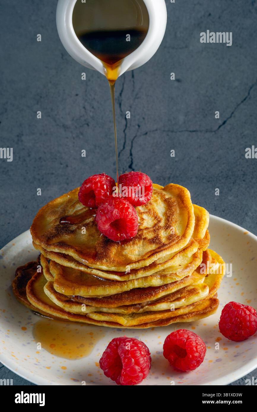 une pile de crêpes à la banane dorée sur une assiette blanche, surmontée de framboises fraîches, comme le sirop d'érable est versé sur eux Banque D'Images