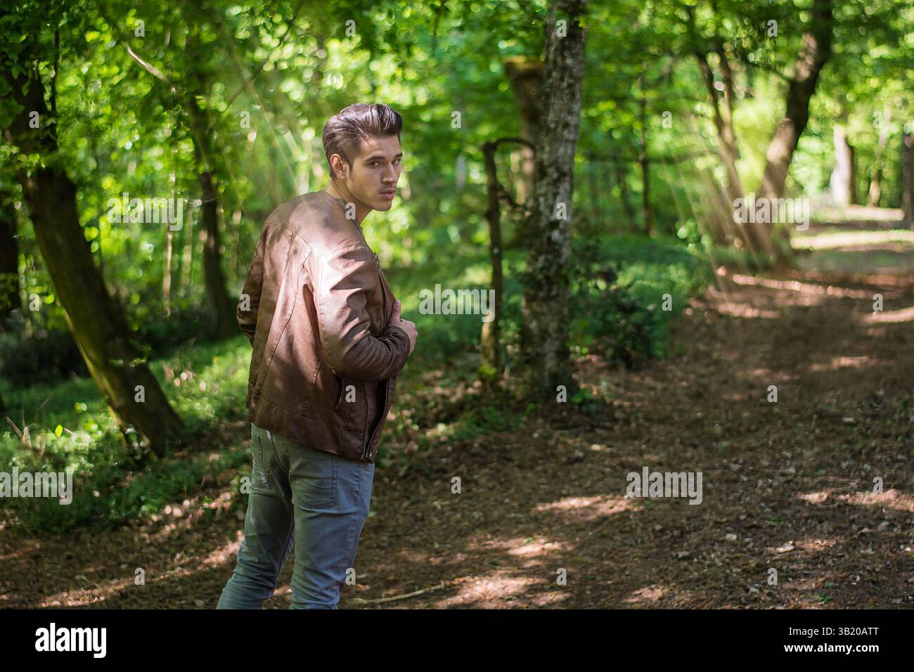 Un jeune homme en tenue décontractée se promène le long d'un chemin de terre dans une forêt verdoyante. La lumière du soleil filtre à travers les arbres, créant une atmosphère sereine alors qu'il regarde en arrière par-dessus son épaule. Banque D'Images