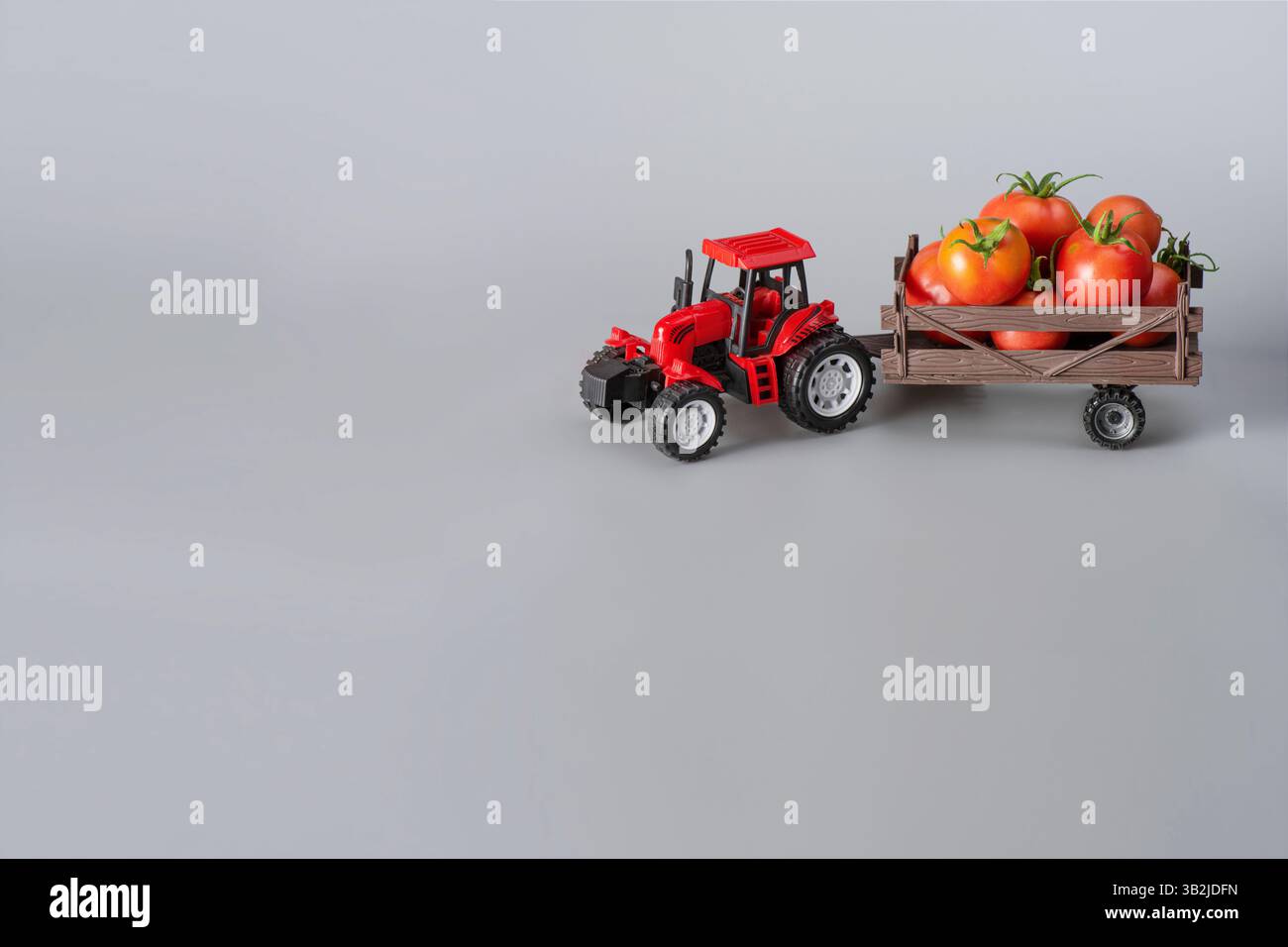 Tracteur jouet rouge tirant une petite remorque en bois chargée de tomates mûres, représentant l'agriculture, l'agriculture, et le voyage de la ferme à la table sur un s. Banque D'Images