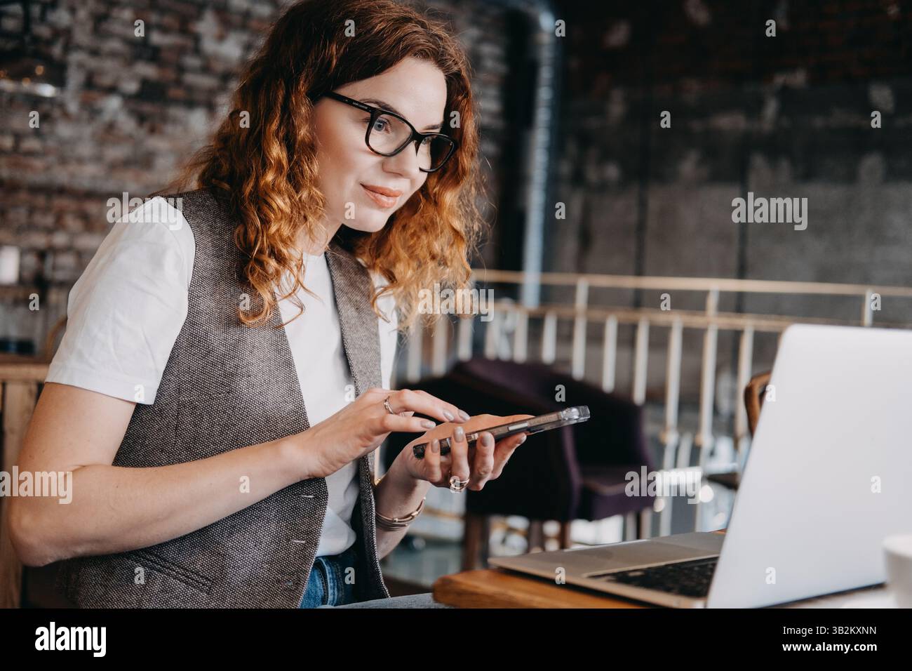 Une jeune femme aux cheveux roux frisés vérifie son smartphone tout en travaillant sur son ordinateur portable dans un café. Multitâche numérique, flux de travail multiplateforme, à distance Banque D'Images