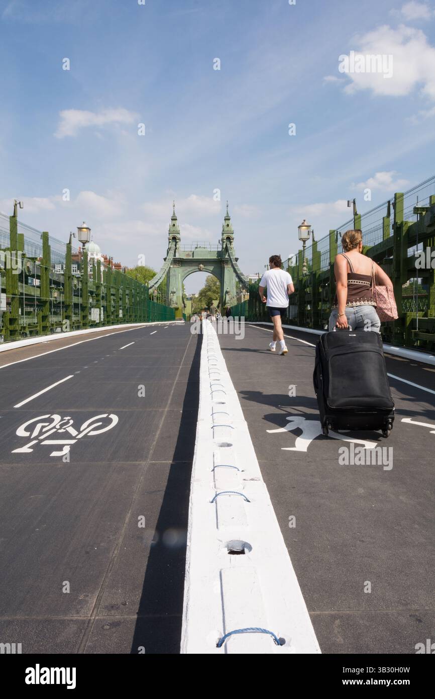 28 avril 2025. Vue arrière d'une jeune femme tirant une grande valise à travers Hammersmith Bridge, qui a maintenant ouvert à nouveau pour les piétons et les cyclistes. Londres, Angleterre, Royaume-Uni Banque D'Images