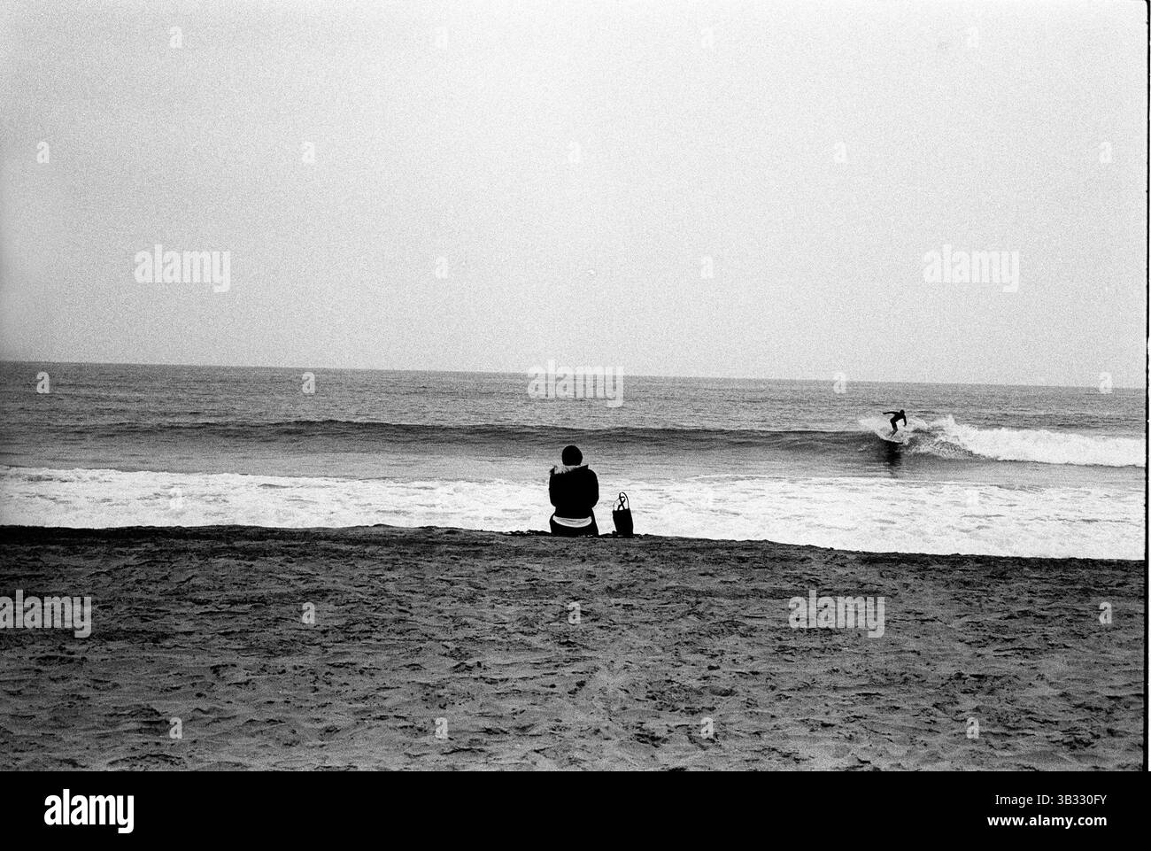 Jan 4, 2016 - South Beach, KwaZulu-Natal, Afrique du Sud - une femme assise sur le sable tandis que d'un surfer rides les vagues. South Beach est une partie de la ville de Durban sans interruption la plus longue étendue de plage de sable. La ville de Durban est sur la côte est de l'Afrique du Sud et les gens ici sont lavés avec de l'eaux chaudes de l'Océan Indien. Au nord de cette étendue de sable sont des plages avec la société hang out cafe. Au sud il y a une jetée avec le quartier haut Moyo's Restaurant à elle est la fin et l'uShaka Marine World et le secteur complexe de la mer et surf clubs de la plage les vesces. Entre Banque D'Images