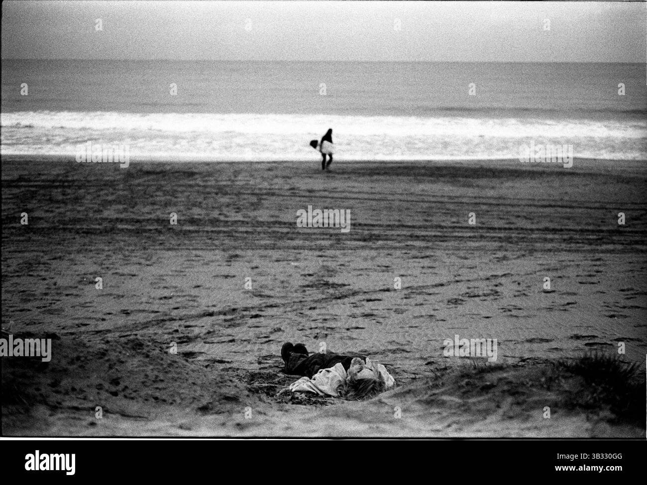 Jan 4, 2016 - South Beach, KwaZulu-Natal, Afrique du Sud - un sans-abri dort dans la lumière des projecteurs de la plage comme un internaute quitte les vagues. South Beach est une partie de la ville de Durban sans interruption la plus longue étendue de plage de sable. La ville de Durban est sur la côte est de l'Afrique du Sud et les gens ici sont lavés avec de l'eaux chaudes de l'Océan Indien. Au nord de cette étendue de sable sont des plages avec la société hang out cafe. Au sud il y a une jetée avec le quartier haut Moyo's Restaurant à elle est la fin et l'uShaka Marine World et le secteur complexe de la mer et surf Banque D'Images
