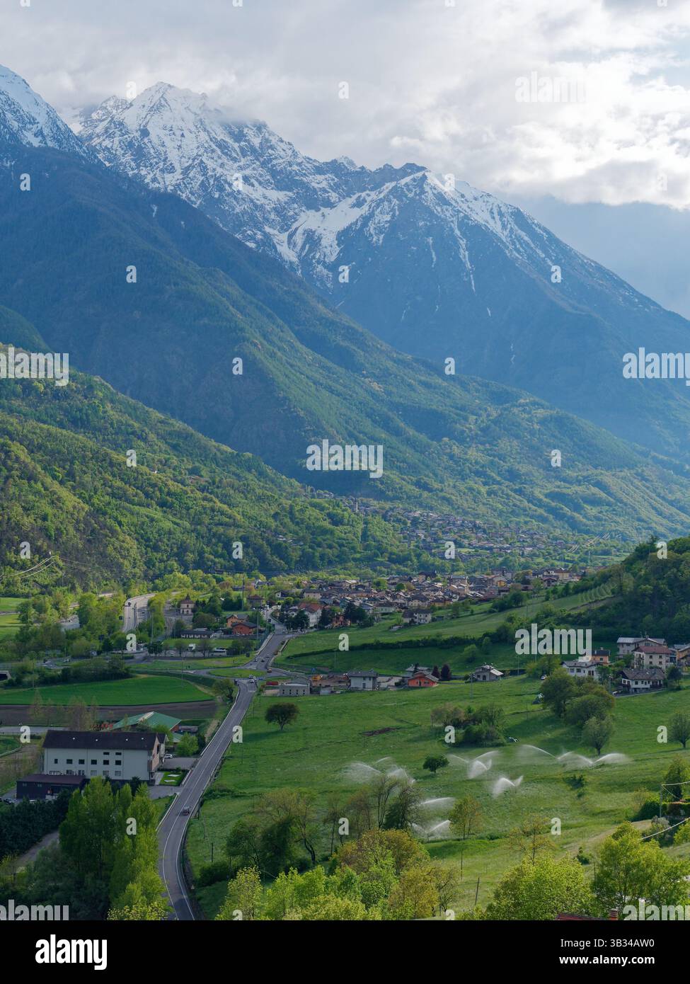 Ville de nus et avec des nuages au-dessus des montagnes enneigées tandis que les arroseurs arrosent un champ en dessous. Région de la vallée d'Aoste, NW Italie. Du 26 au 27 avril 2025 Banque D'Images