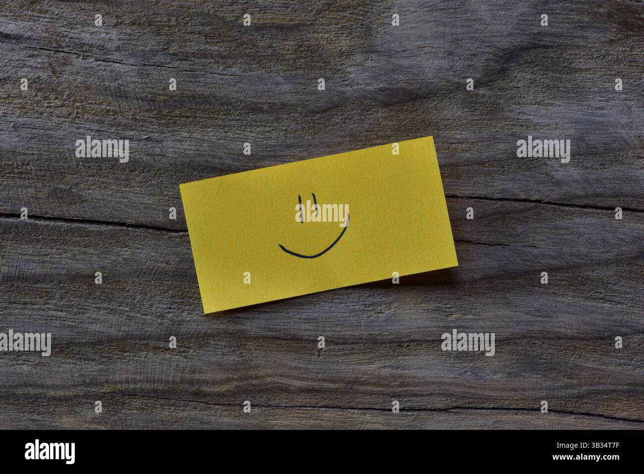 Visage souriant sur note collante jaune sur fond texturé de planche de bois. Un stylo dessine un visage souriant sur du papier à lettres. Banque D'Images