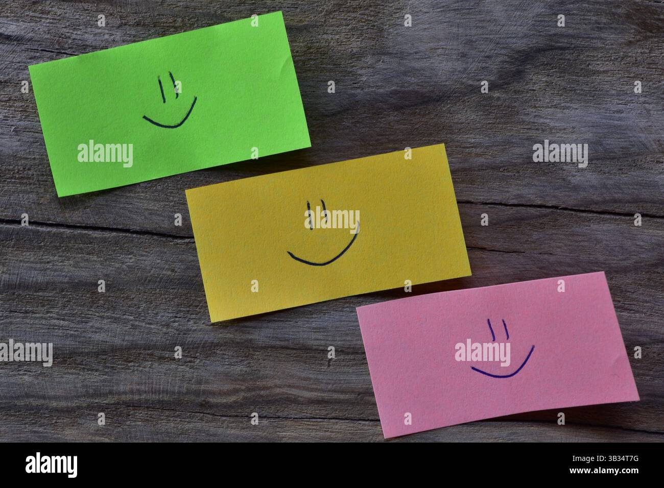 Visage souriant sur note collante verte, jaune et rose sur fond texturé de planche de bois. Un stylo dessine un visage souriant sur du papier à notes. Banque D'Images